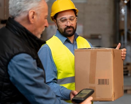 Dos trabajadores en un almacén, uno escanea una caja con un código de barras mientras el otro la sostiene